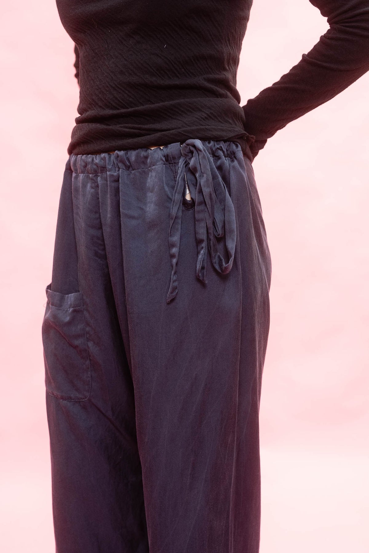 A person wearing loose navy drawstring pants with side cargo pockets, paired with a black ribbed long-sleeve top, against a soft pink background.