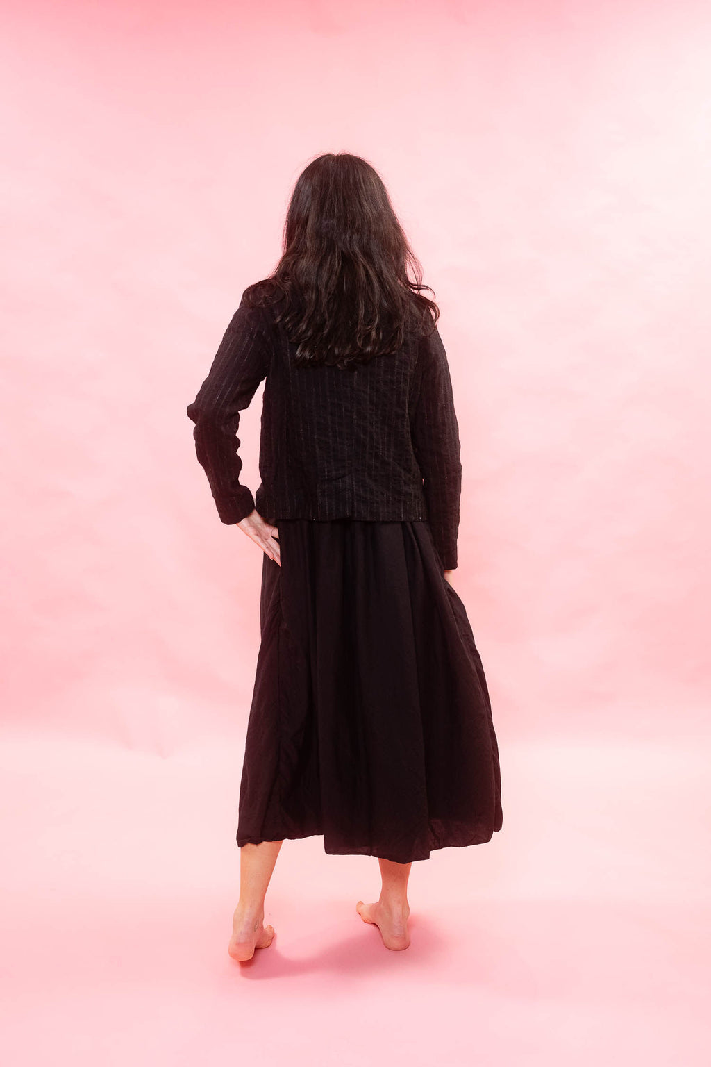 A woman is shown from behind wearing a black textured jacket and a long black skirt, standing against a solid pink studio backdrop.