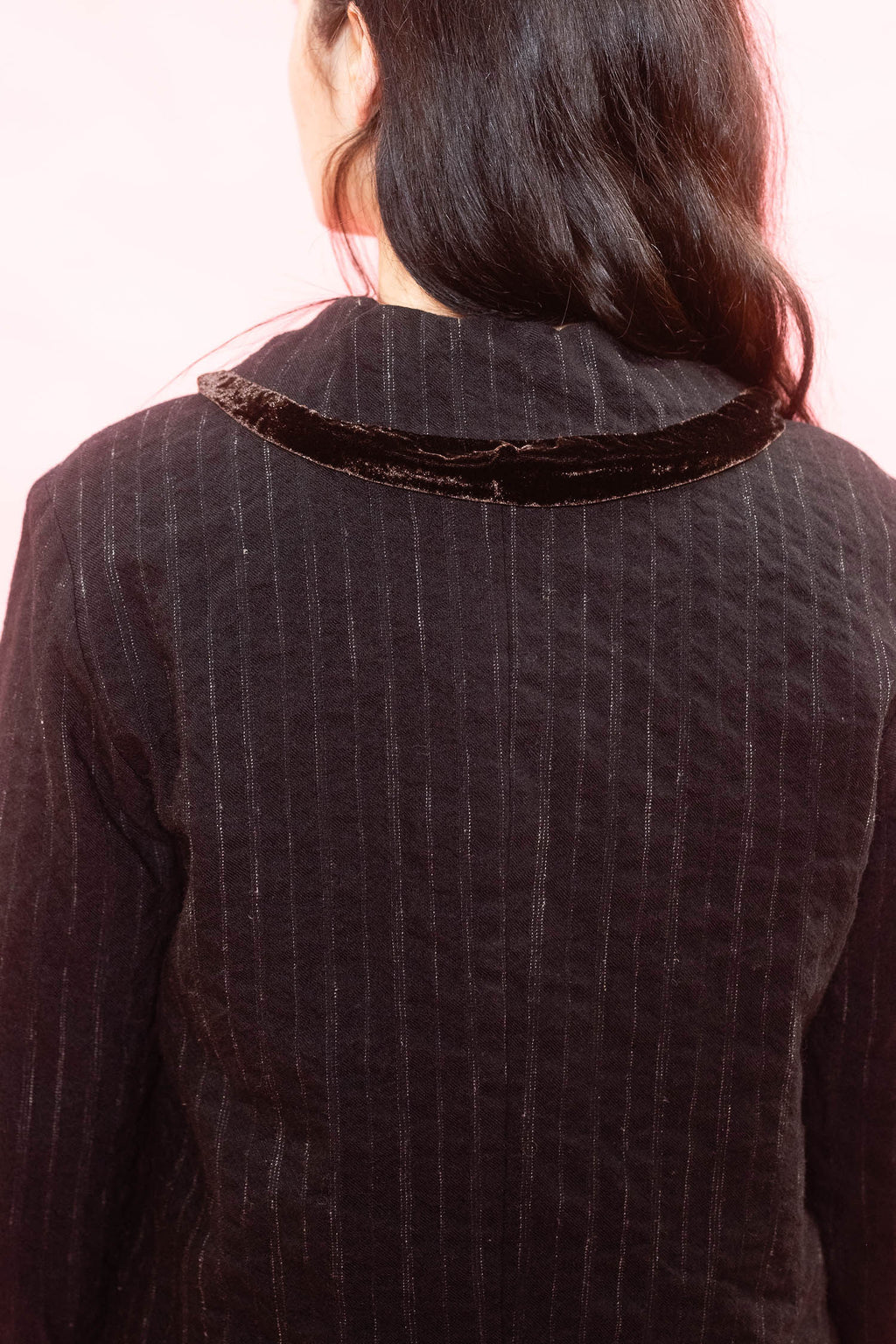 Back view of a person wearing a black pinstriped jacket with a contrasting dark brown velvet collar, set against a solid light pink background.