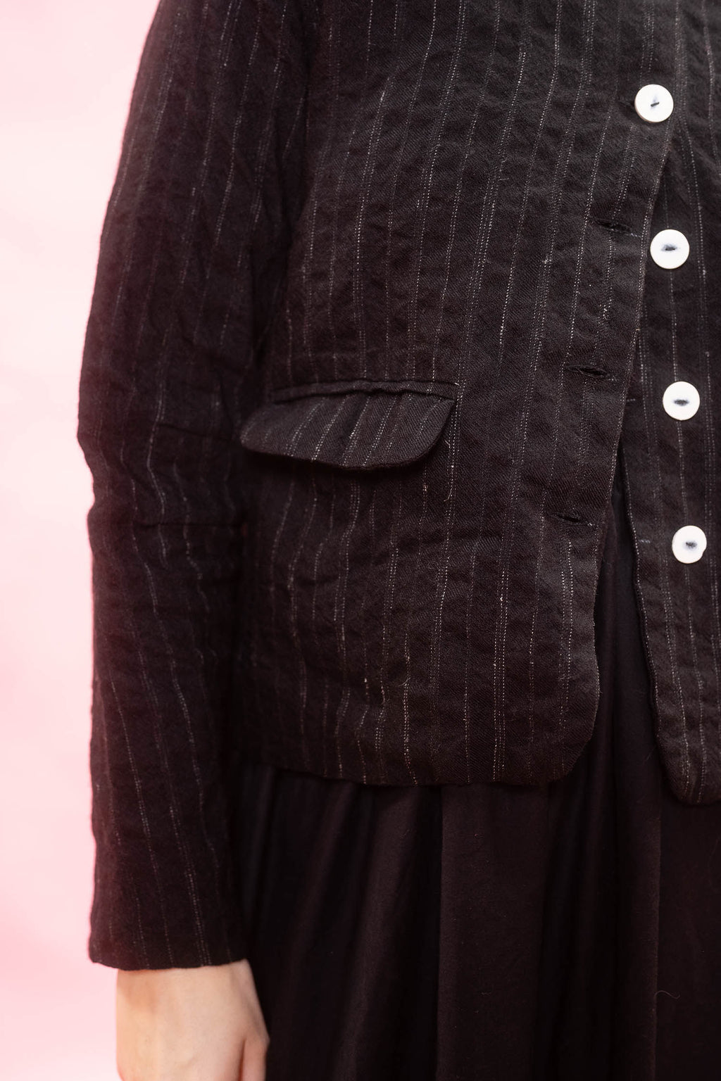 Close-up of a person wearing a black textured jacket with vertical pinstripes, white buttons, and a flap pocket, paired with a dark skirt against a soft pink background.