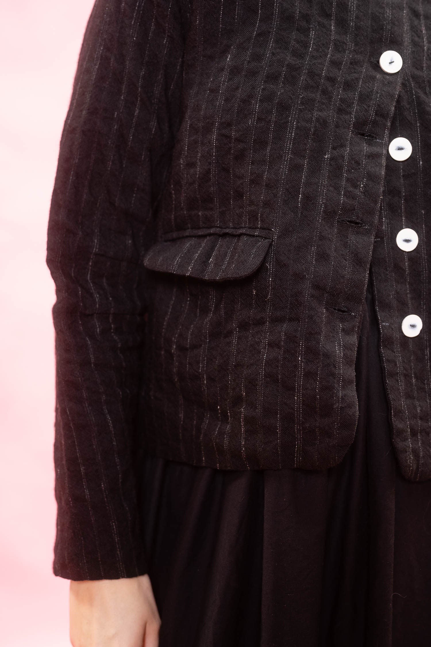 Close-up of a person wearing a black textured jacket with vertical pinstripes, white buttons, and a flap pocket, paired with a dark skirt against a soft pink background.