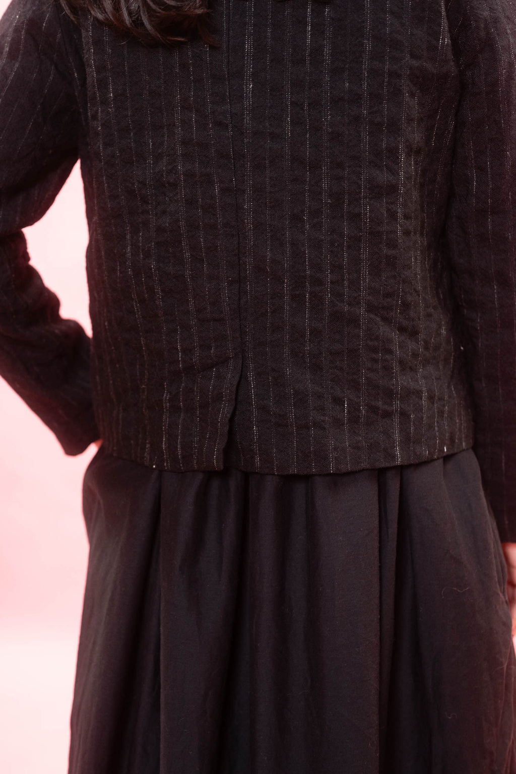 Close-up of a person wearing a dark charcoal pinstriped jacket with a pleated waist and a dark skirt, set against a soft pink background.