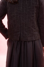Close-up of a person wearing a dark charcoal pinstriped jacket with a pleated waist and a dark skirt, set against a soft pink background.