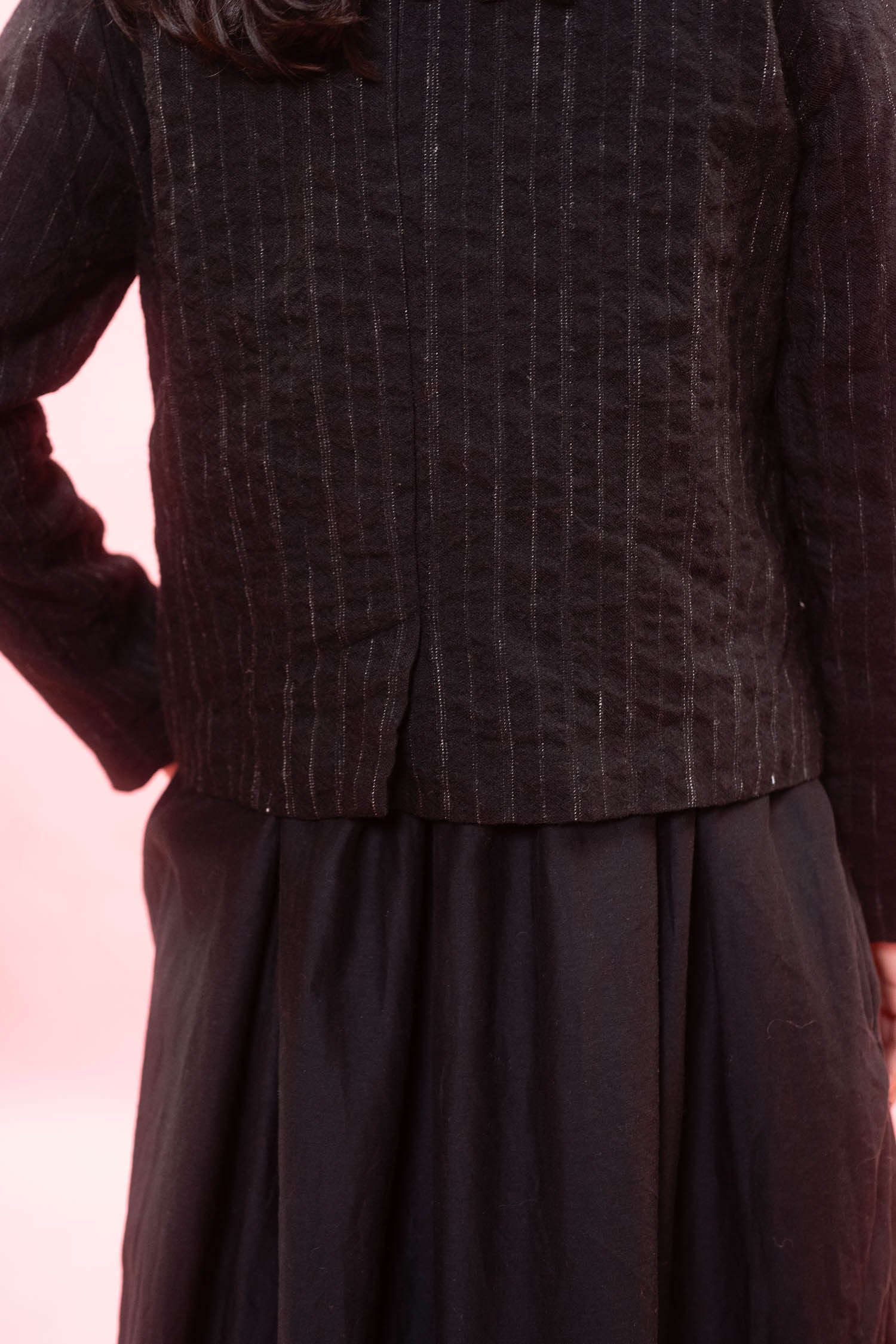 Close-up of a person wearing a dark charcoal pinstriped jacket with a pleated waist and a dark skirt, set against a soft pink background.