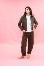 A woman stands barefoot against a solid pink studio background, wearing a dirt green cotton patina jacket and matching trousers over a white collared shirt.