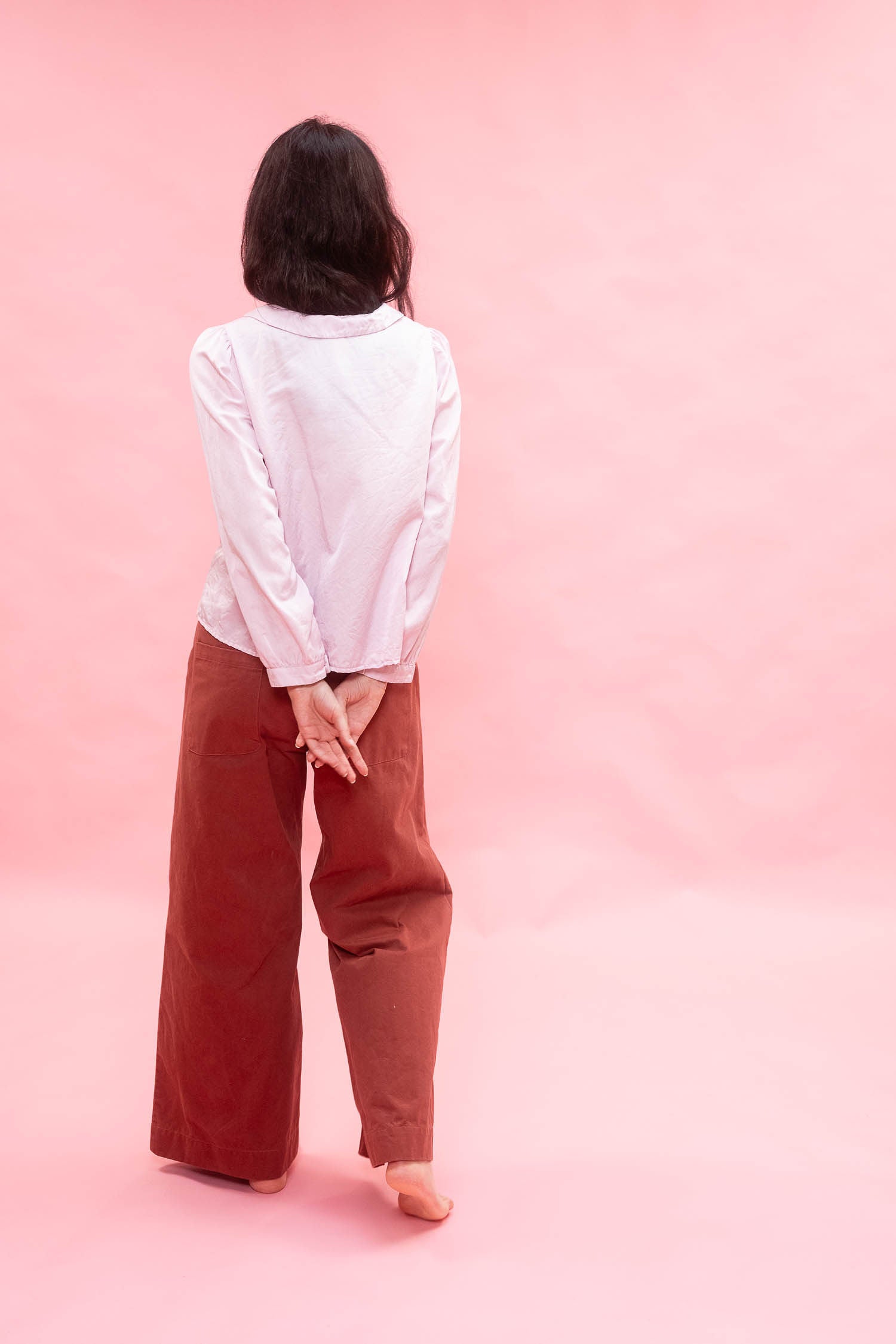 A woman standing with her back to the camera, wearing a pink long-sleeve button-up shirt made from silk cotton satin and wide-leg rust-brown pants against a solid pink studio background.

