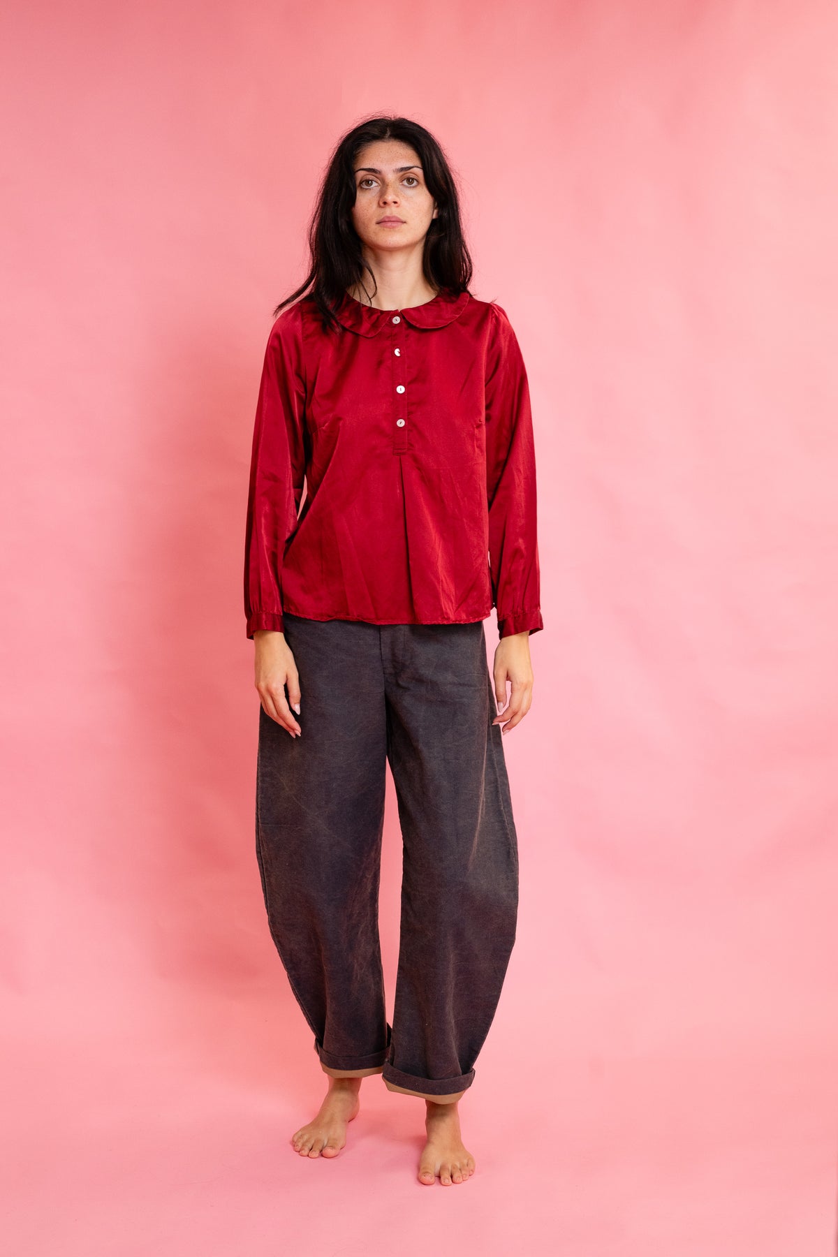 A woman stands barefoot against a solid pink studio backdrop, wearing a long-sleeve red button-front top with a wide collar and loose dark navy pants.