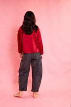 A model standing with their back to the camera, wearing a bright red long-sleeve top and loose dark navy pants, set against a solid pink studio background.