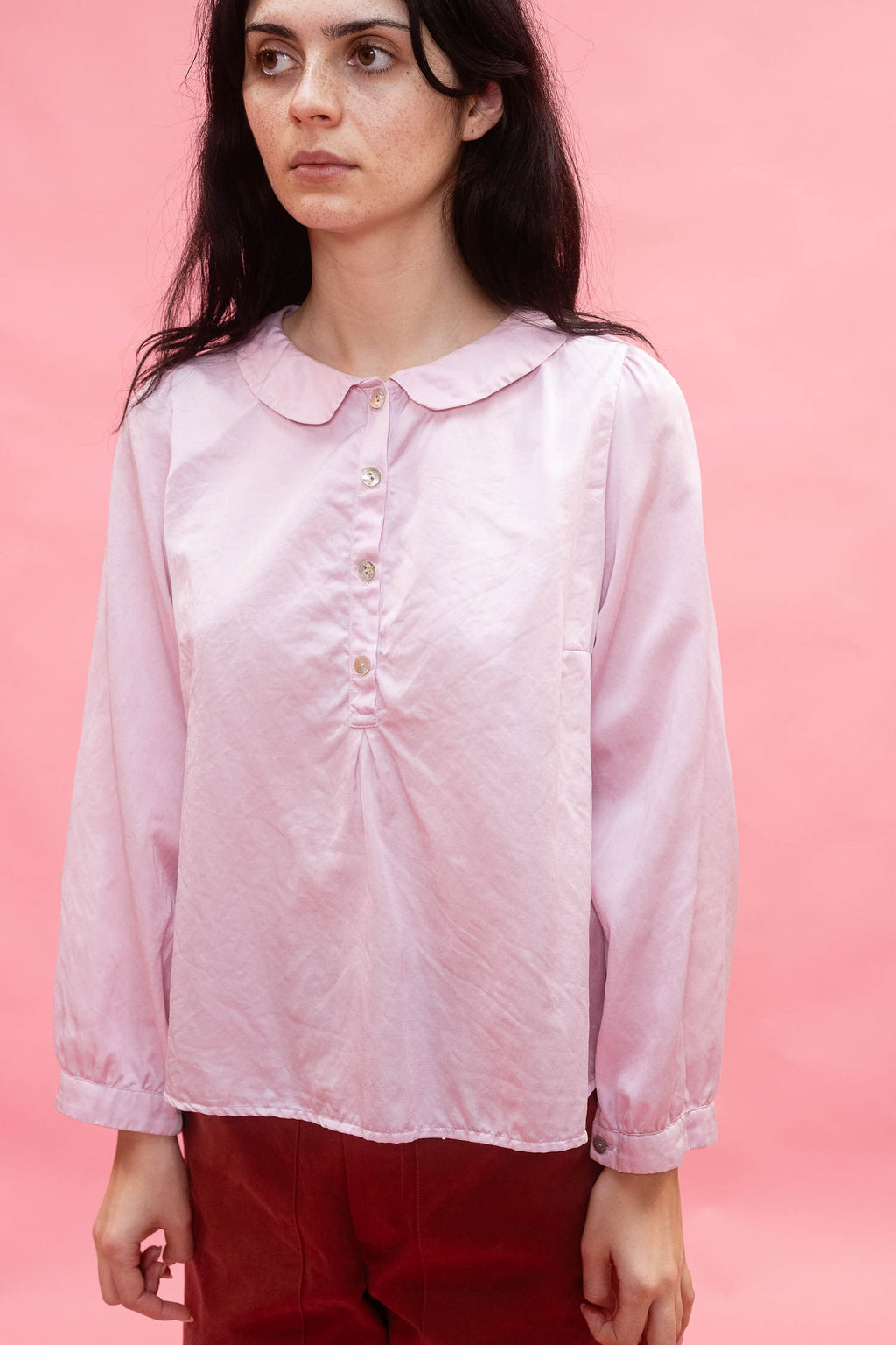 A woman wearing a light pink long-sleeve blouse with a round Peter Pan collar and button placket, made with silk cotton satin and paired with dark red pants, set against a solid pink background.
