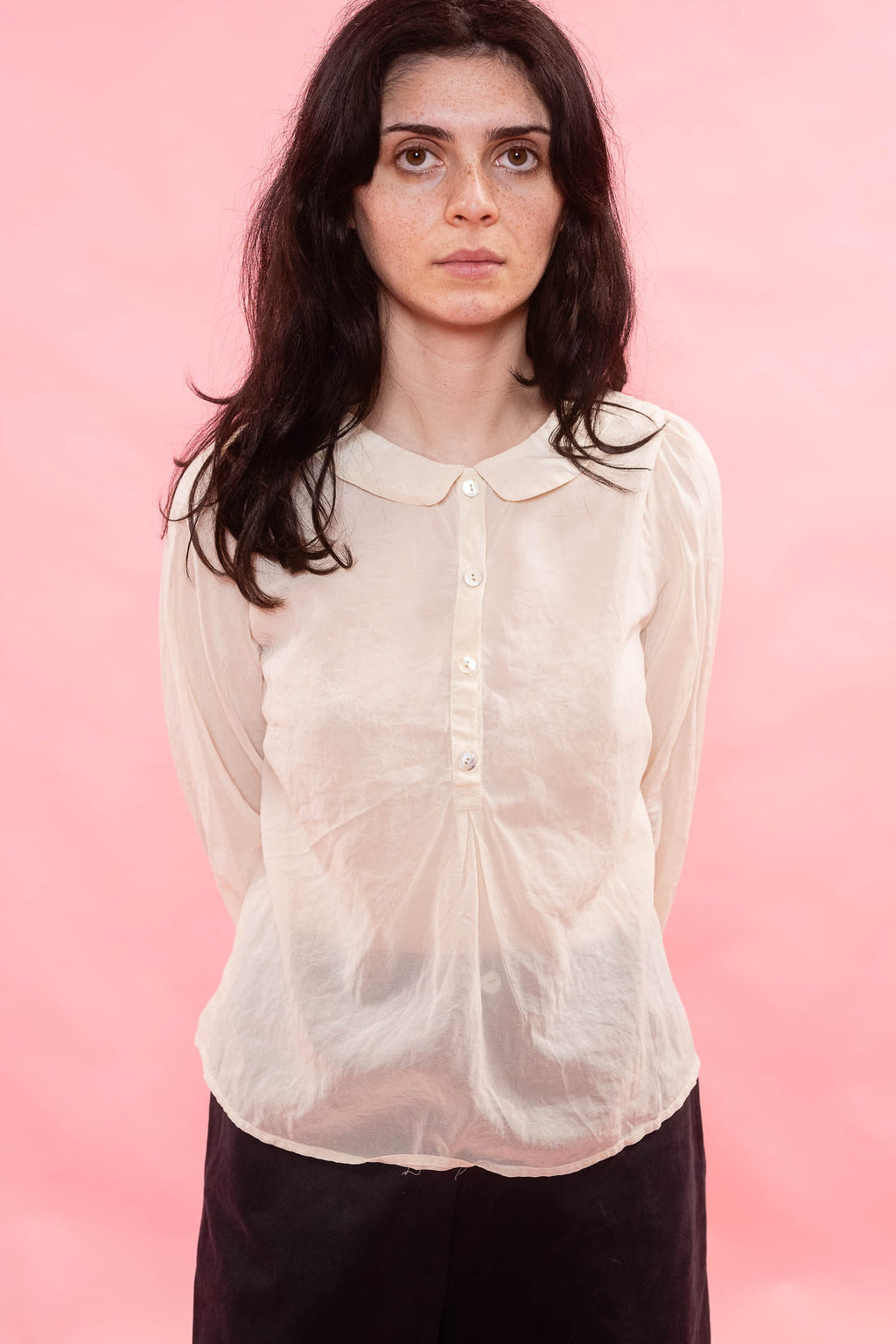 A woman with dark hair wearing a semi-sheer, cream organdy long-sleeve blouse with a Peter Pan collar and front buttons, paired with dark trousers, set against a solid pink background.
