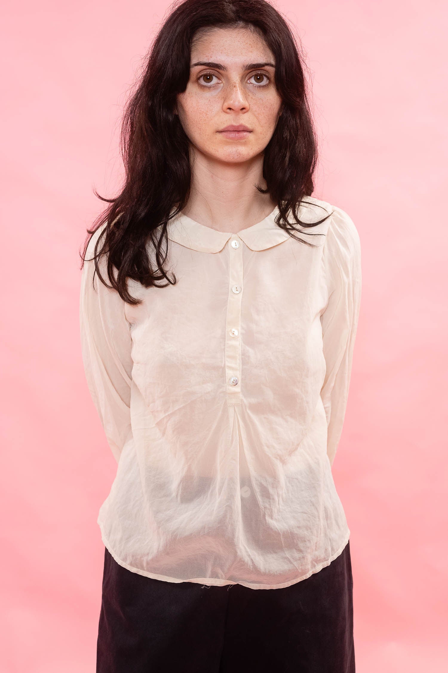 A woman with dark hair wearing a semi-sheer, cream organdy long-sleeve blouse with a Peter Pan collar and front buttons, paired with dark trousers, set against a solid pink background.
