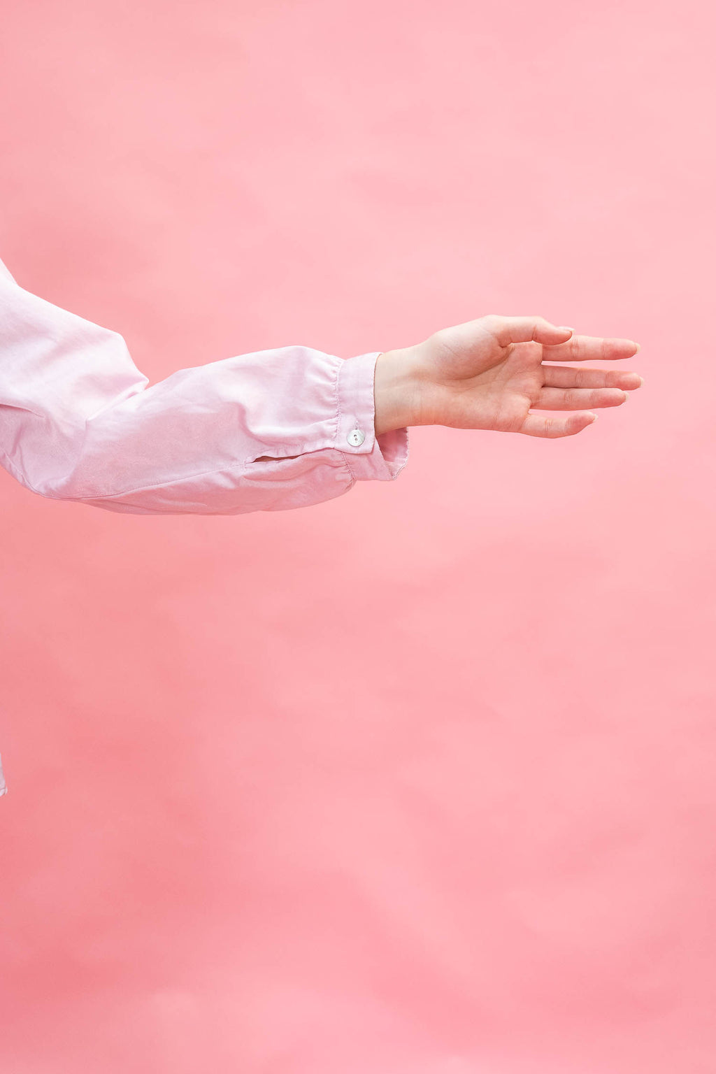 A person's hand and forearm wearing a light pink long-sleeve button-up shirt, extending toward the right against a solid pink background.
