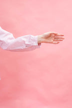 A person's hand and forearm wearing a light pink long-sleeve button-up shirt, extending toward the right against a solid pink background.
