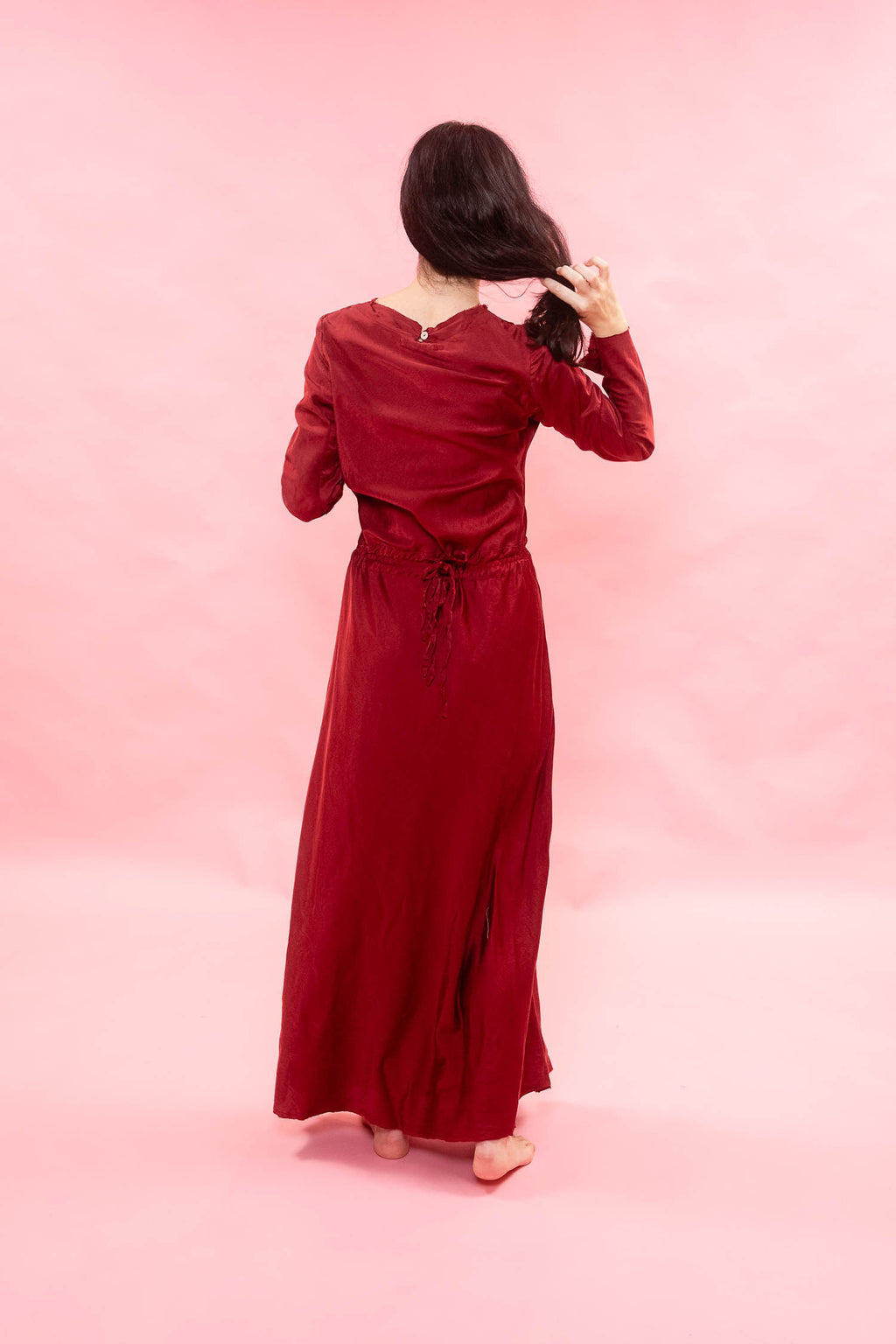 A woman with long dark hair is shown from behind wearing a long-sleeved, floor-length burgundy dress with a tie waist, standing against a solid pink studio backdrop.