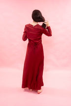 A woman with long dark hair is shown from behind wearing a long-sleeved, floor-length burgundy dress with a tie waist, standing against a solid pink studio backdrop.