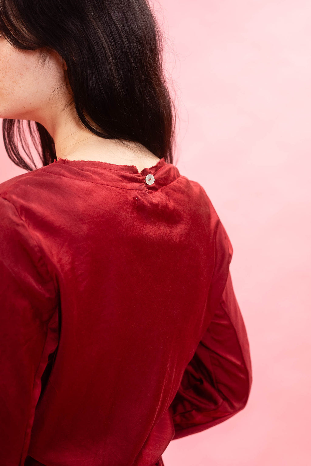 A close-up back view of a person wearing a long-sleeve red velvet top against a solid pink background.