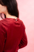 A close-up back view of a person wearing a long-sleeve red velvet top against a solid pink background.