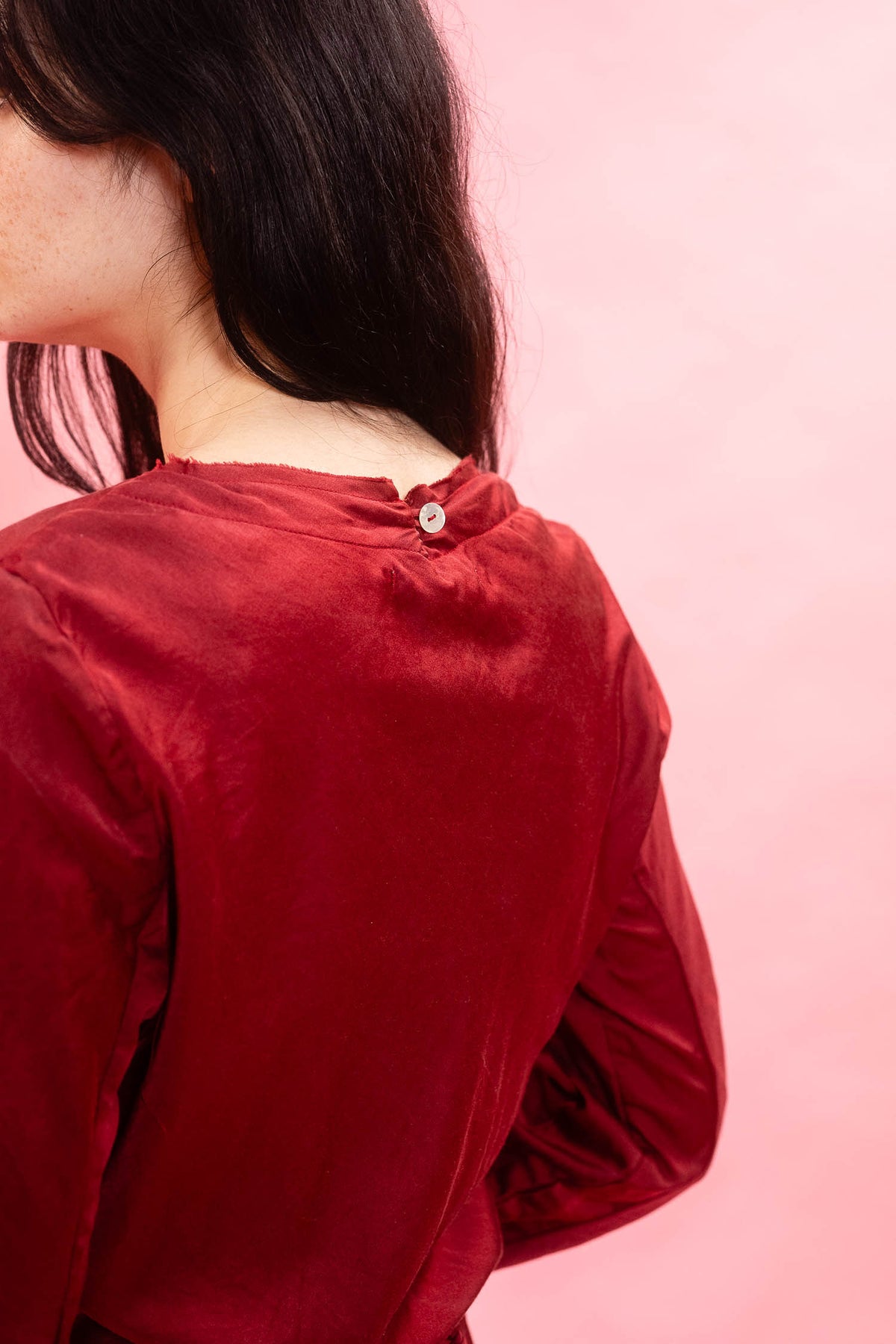A close-up back view of a person wearing a long-sleeve red velvet top against a solid pink background.