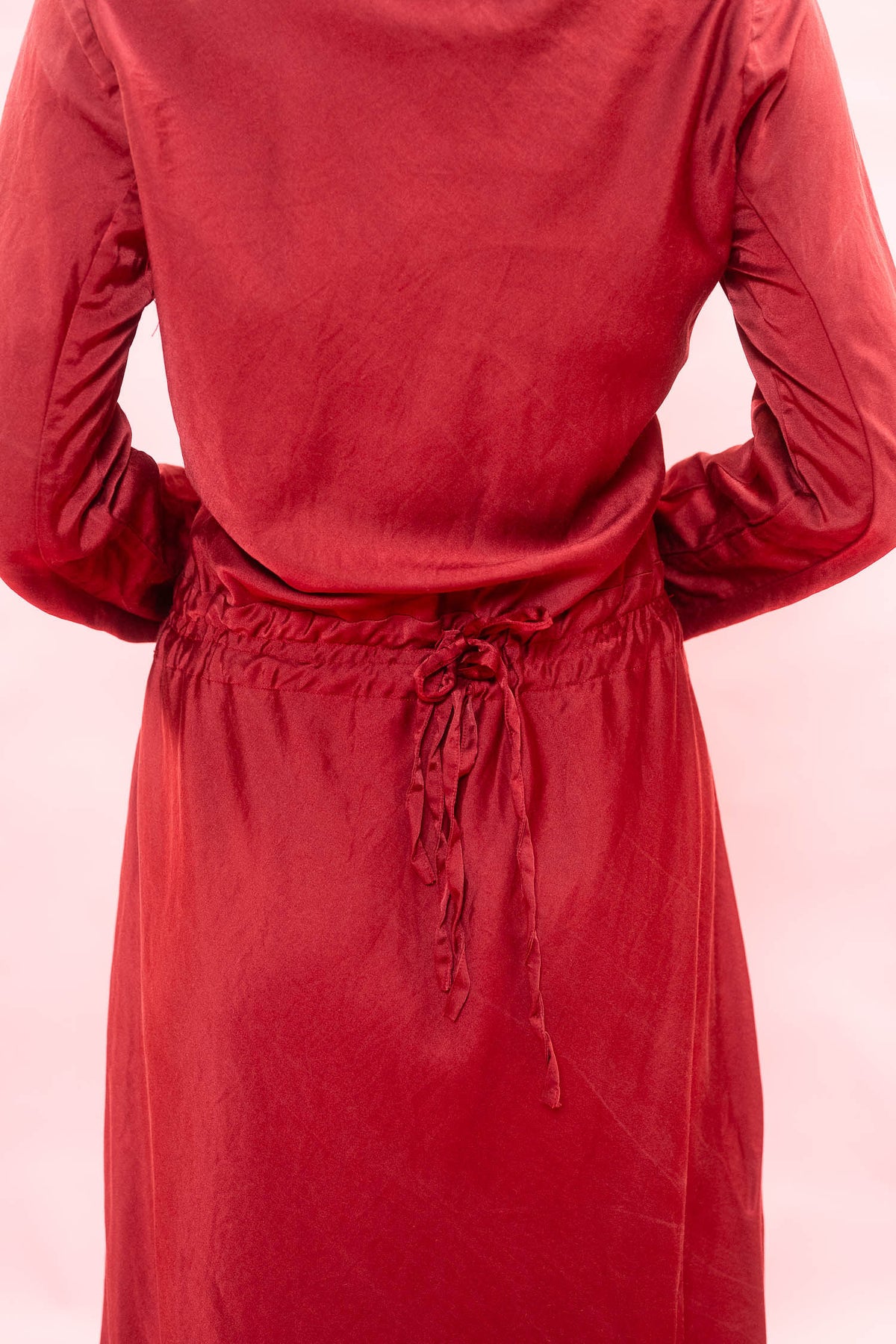 A close-up back view of a person wearing a long-sleeve red dress with a drawstring waist, set against a solid light pink background.