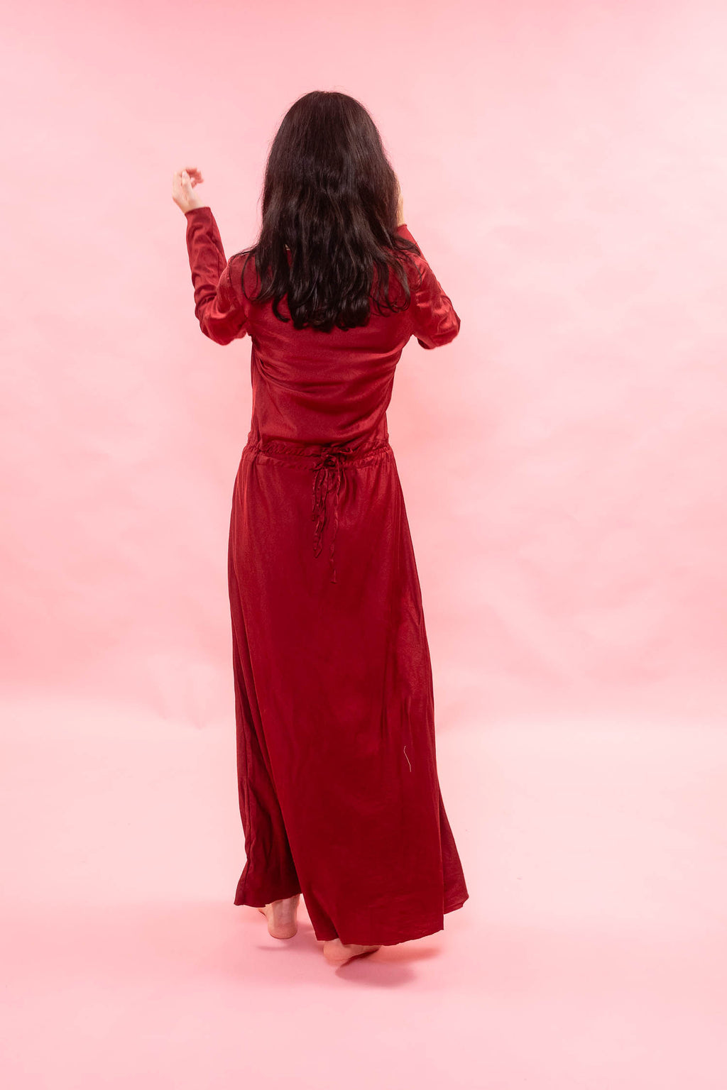 A woman viewed from behind wearing a long-sleeved, floor-length burgundy red dress with a tie waist, standing against a solid pink backdrop.