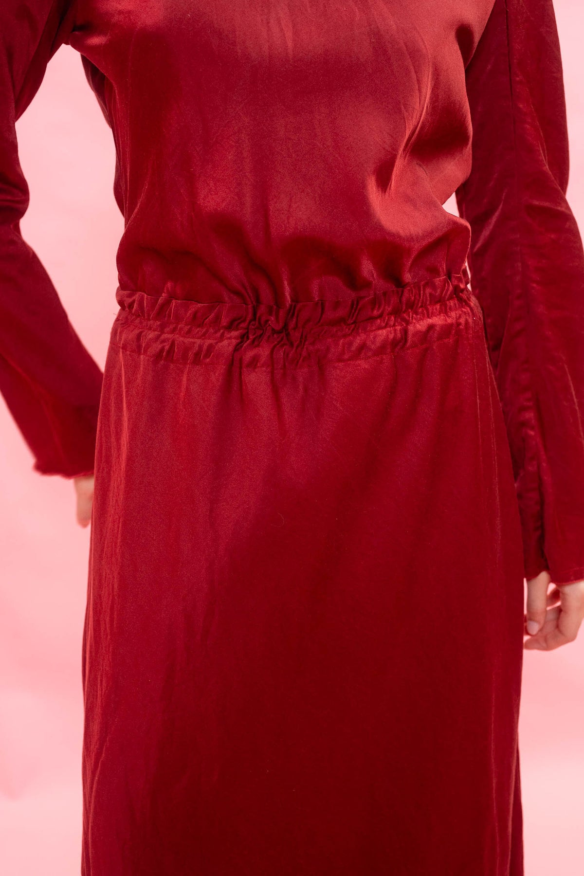 A close-up of a model wearing a long-sleeved red dress with a gathered elastic waist, set against a solid pink background.