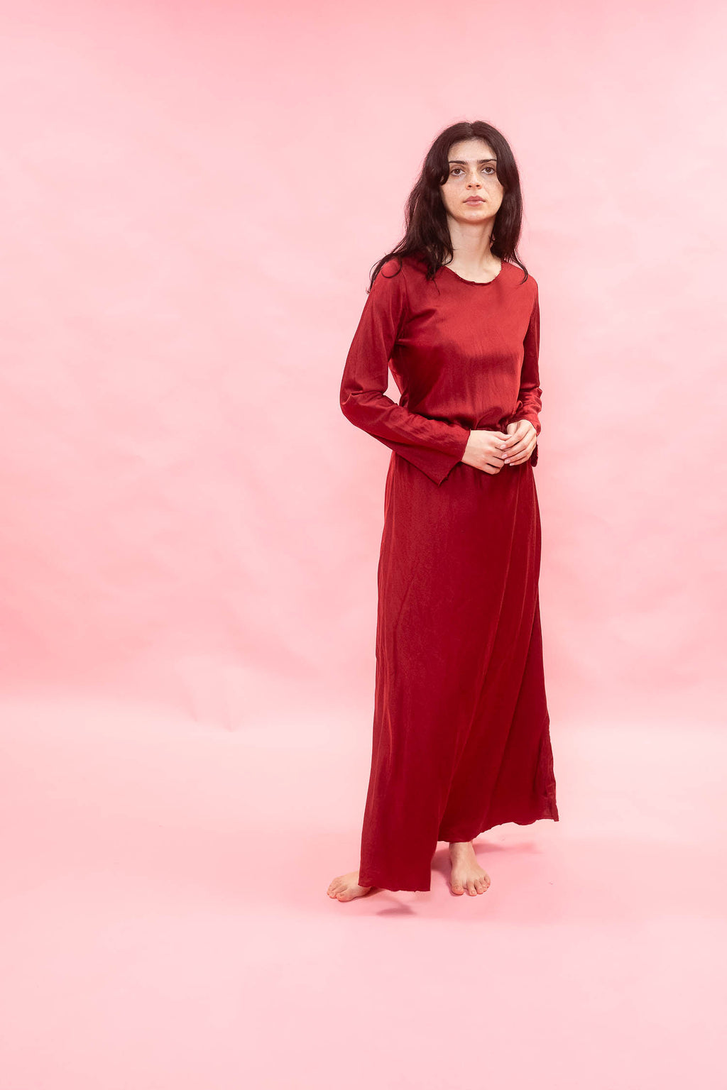 A woman stands barefoot against a solid pink studio backdrop, wearing a long-sleeved, floor-length red dress with a gathered waist and flowing skirt.