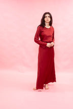 A woman stands barefoot against a solid pink studio backdrop, wearing a long-sleeved, floor-length red dress with a gathered waist and flowing skirt.