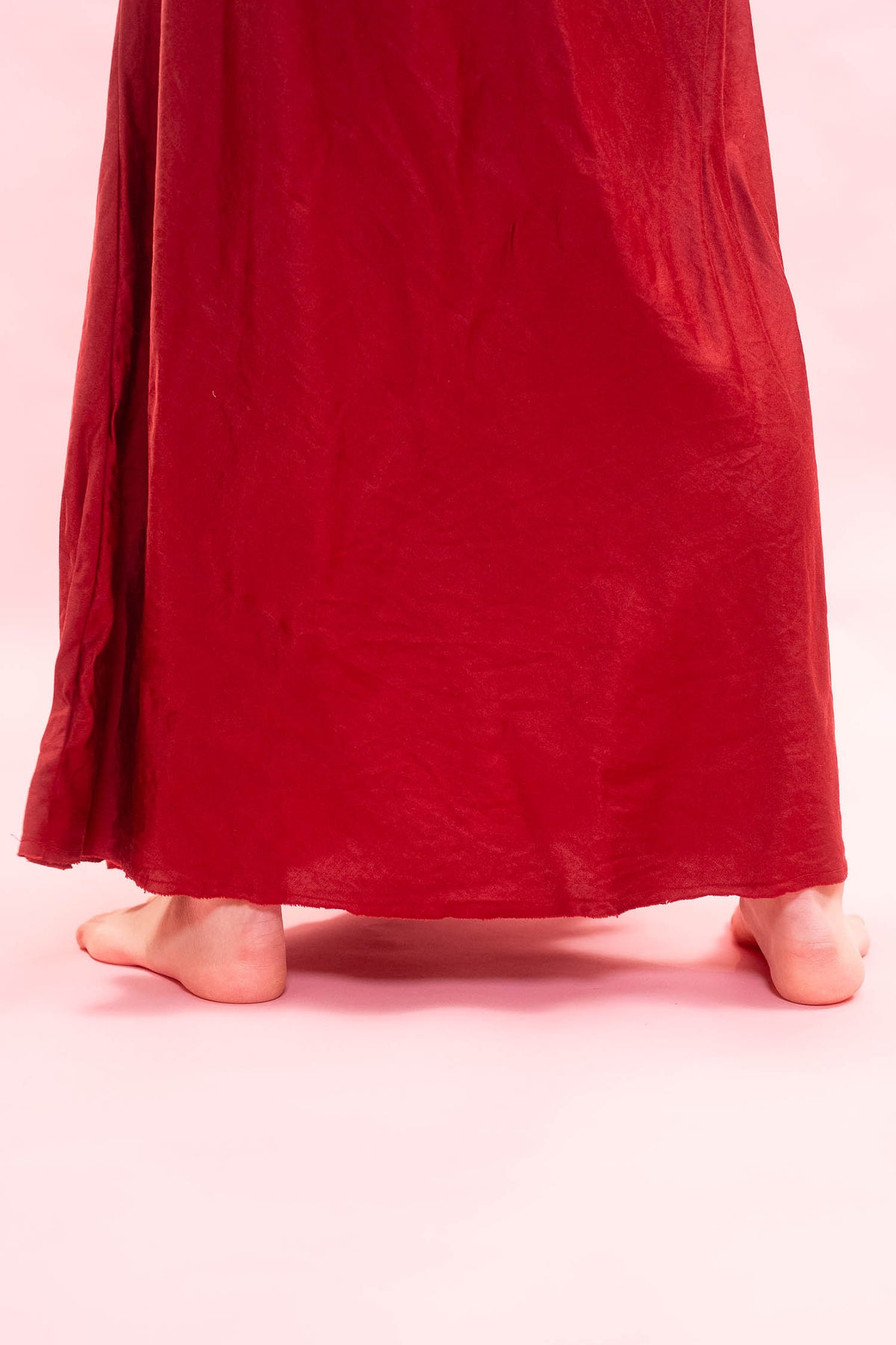 A person wearing a long, flowing red skirt or dress, shown from the back with bare feet against a solid light pink background.