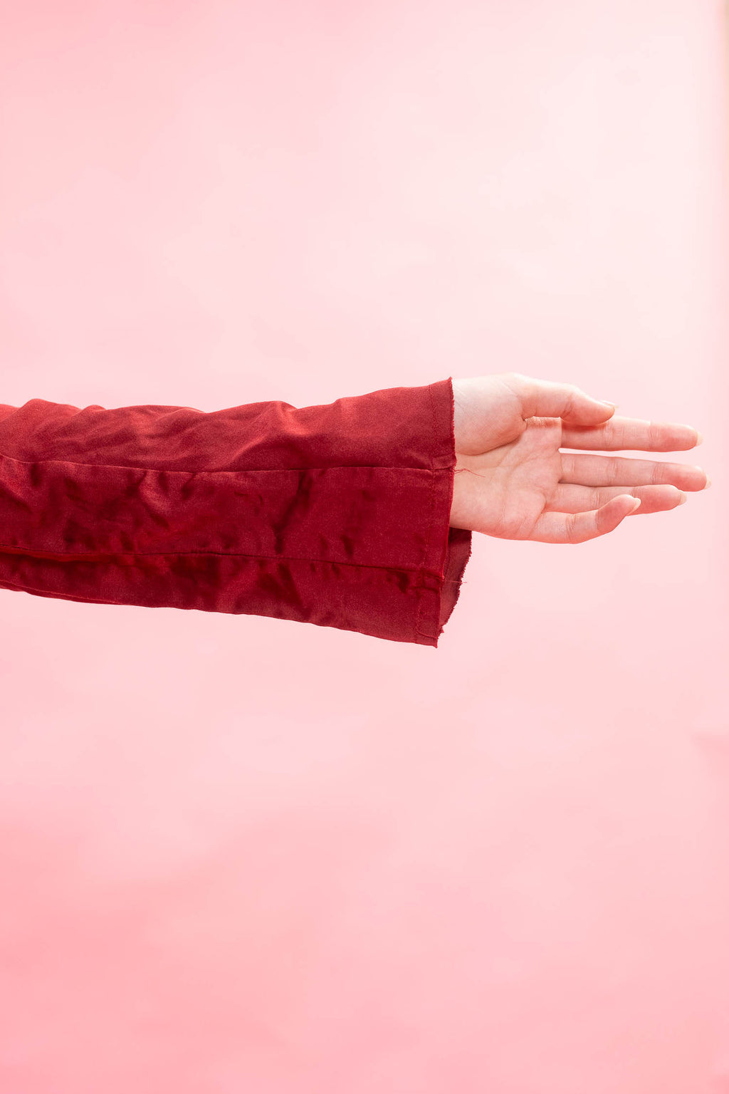 A close-up of a person's hand and forearm wearing a textured, deep red velvet or satin sleeve, set against a solid light pink background.