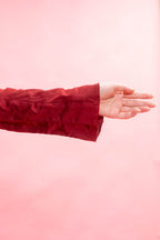 A close-up of a person's hand and forearm wearing a textured, deep red velvet or satin sleeve, set against a solid light pink background.