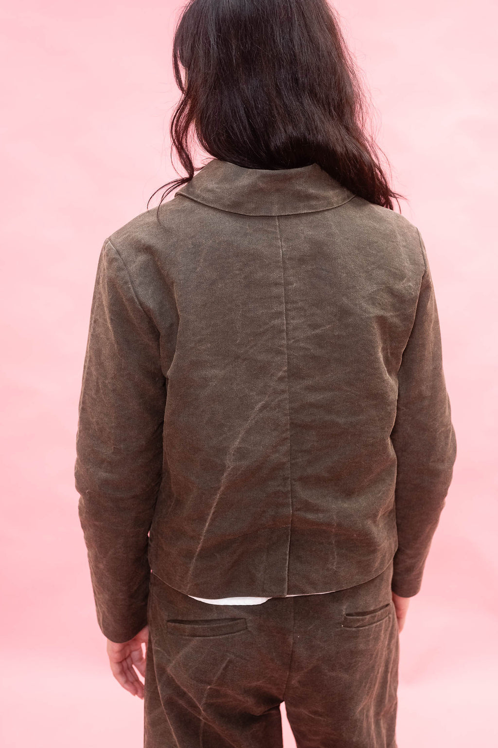 A person shown from the back wearing a dark green cropped jacket and matching pants against a solid pink background.
