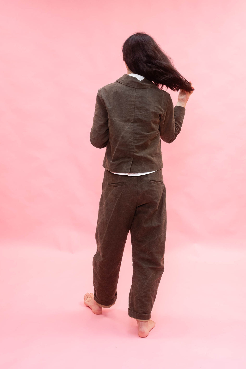 A woman is shown from behind wearing a dark green cotton jacket and matching trousers, standing against a solid pink studio background.