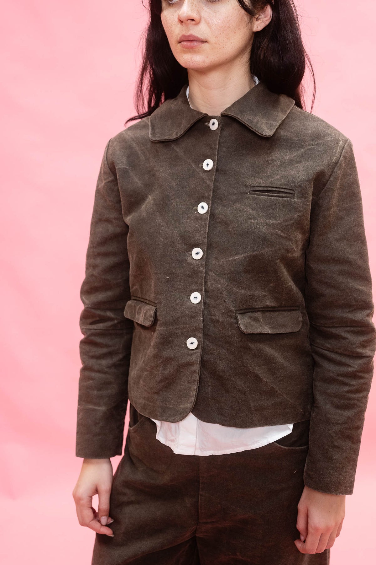A model wearing a dark green cotton jacket with white buttons, styled with matching green trousers and a white shirt, set against a solid pink background.