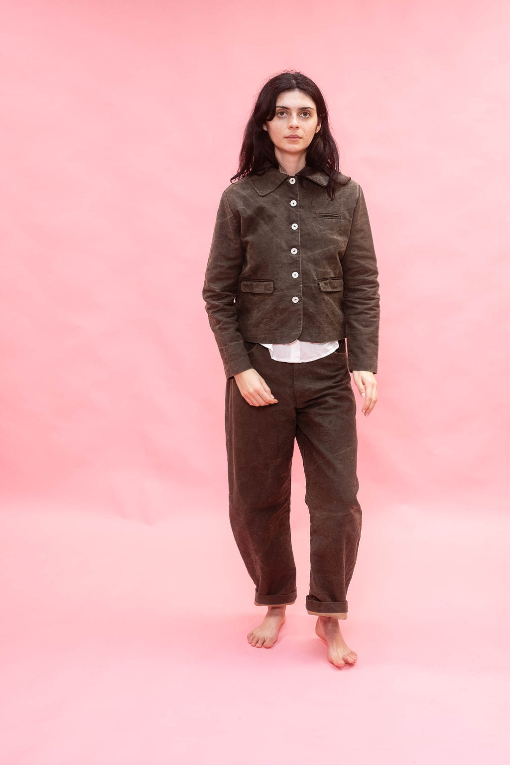A woman stands barefoot against a solid pink studio backdrop, wearing a dark green cotton button-front jacket over a white undershirt and matching dark green cotton pants.