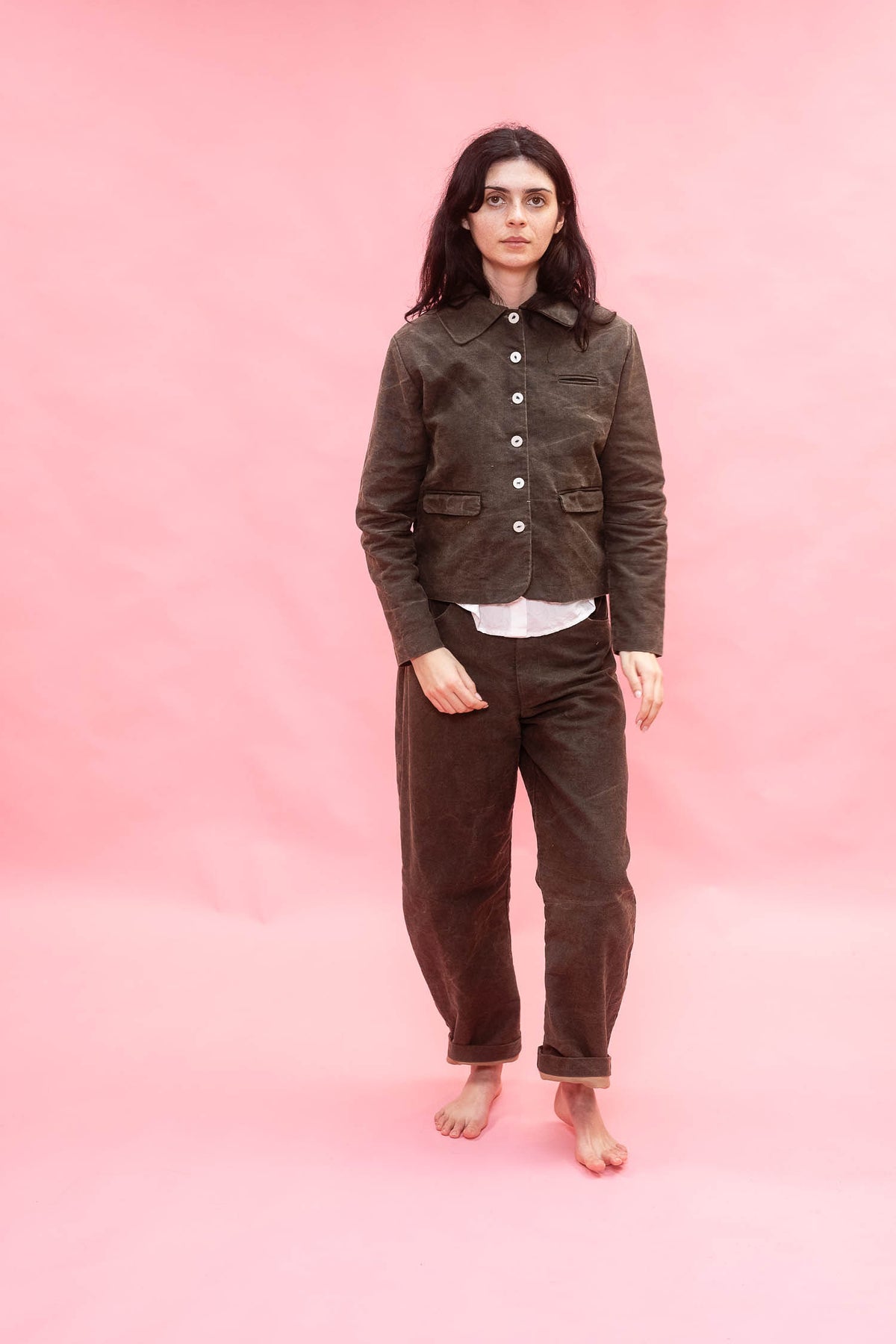 A woman stands barefoot against a solid pink studio backdrop, wearing a dark green cotton button-front jacket over a white undershirt and matching dark green cotton pants.