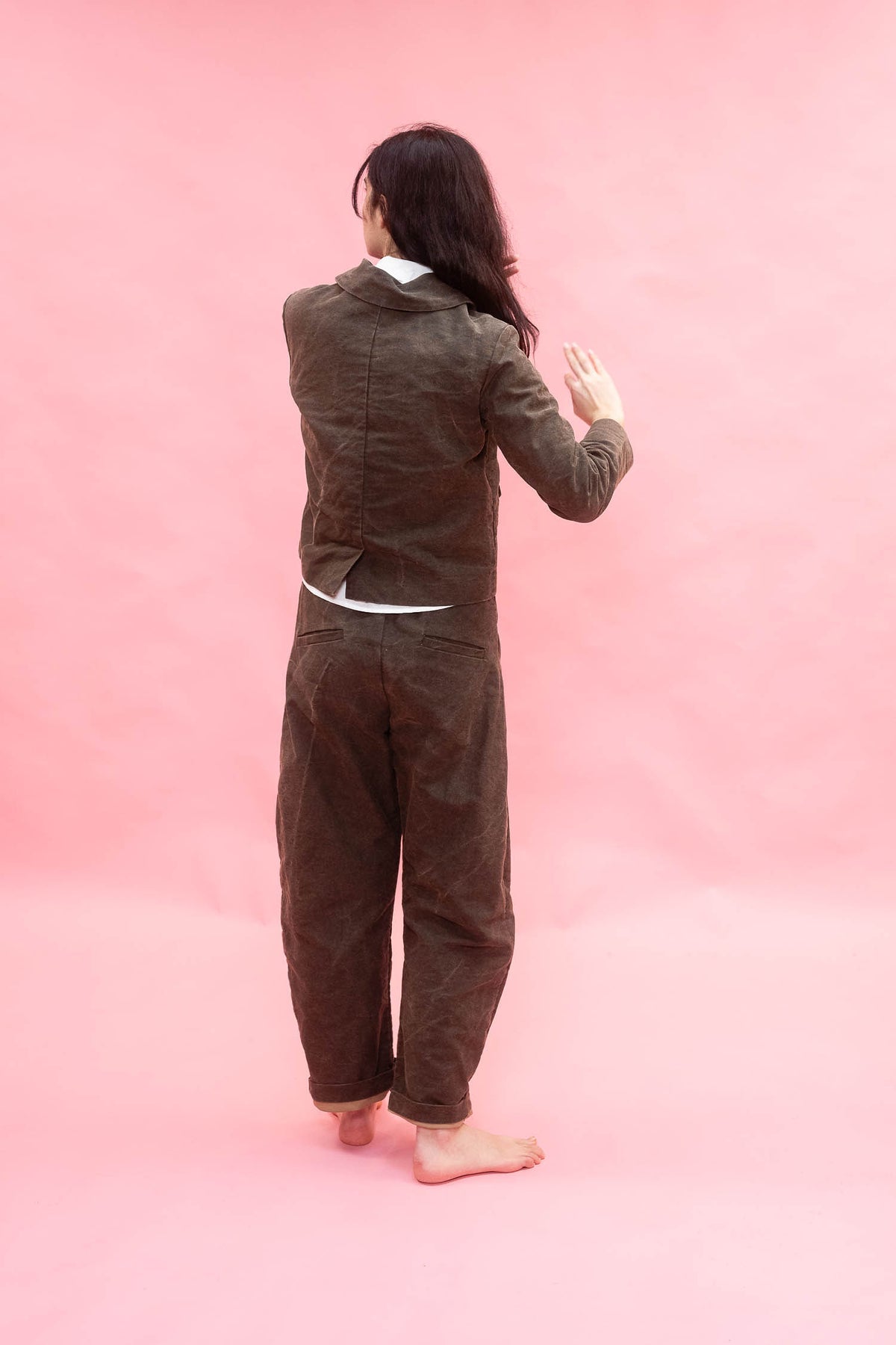A person standing barefoot against a solid pink backdrop, viewed from behind, wearing a dark green cotton jacket and matching pants.