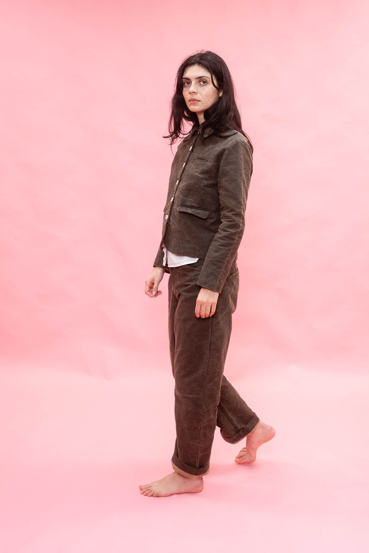 A woman stands barefoot against a solid pink studio backdrop, wearing a dark green cotton  jacket and matching wide-leg trousers over a white undershirt.