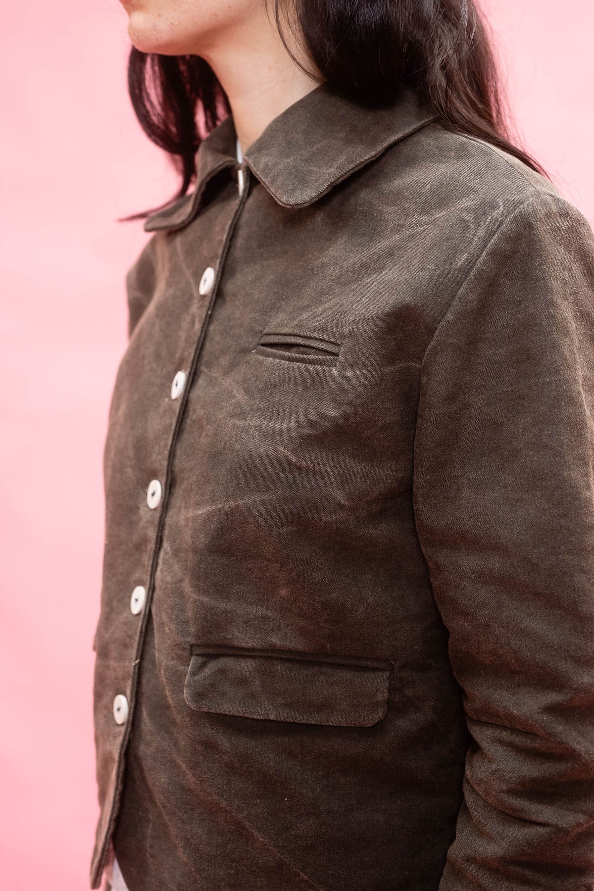 A close-up side view of a person wearing a dark green cotton canvas utility jacket against a solid pink background.