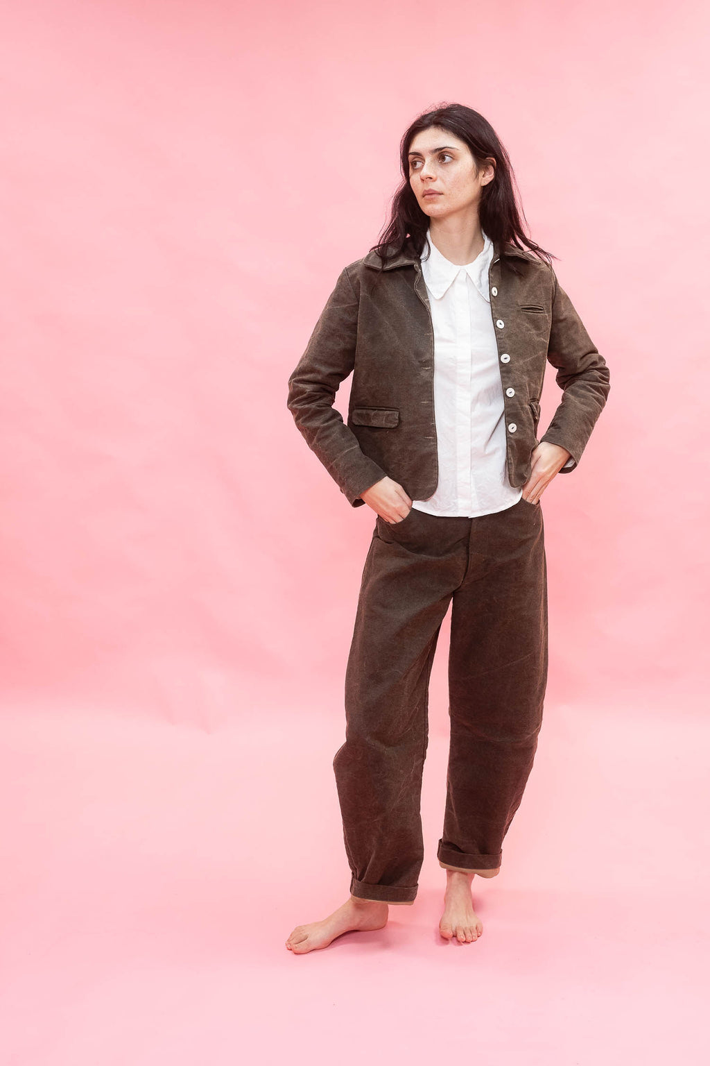 A woman stands barefoot against a solid pink studio background, wearing a dark green cotton jacket and matching trousers over a white collared shirt.