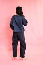 A person standing with their back to the camera, wearing a dark navy blue satin-like long-sleeve top and matching dark pants, set against a solid pink studio background.