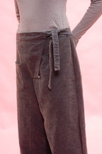 A close-up of a person wearing wide-leg dark navy corduroy pants with a tie waist and side pocket, paired with a light beige long-sleeve top against a soft pink background.