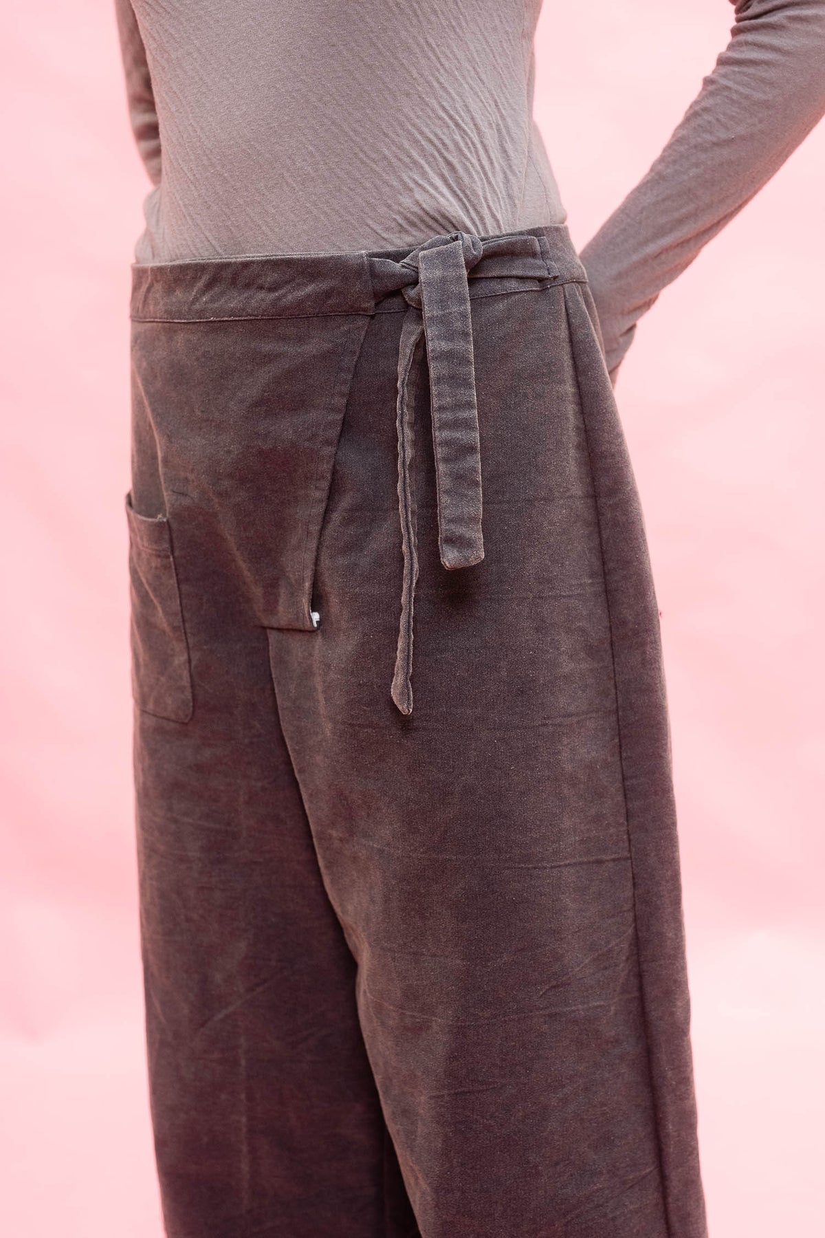 A close-up of a person wearing wide-leg dark navy corduroy pants with a tie waist and side pocket, paired with a light beige long-sleeve top against a soft pink background.