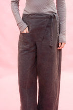 A person wearing dark na wide-leg linen pants with a tie waist and side pockets, paired with a gray ribbed long-sleeve top, set against a solid pink background.y