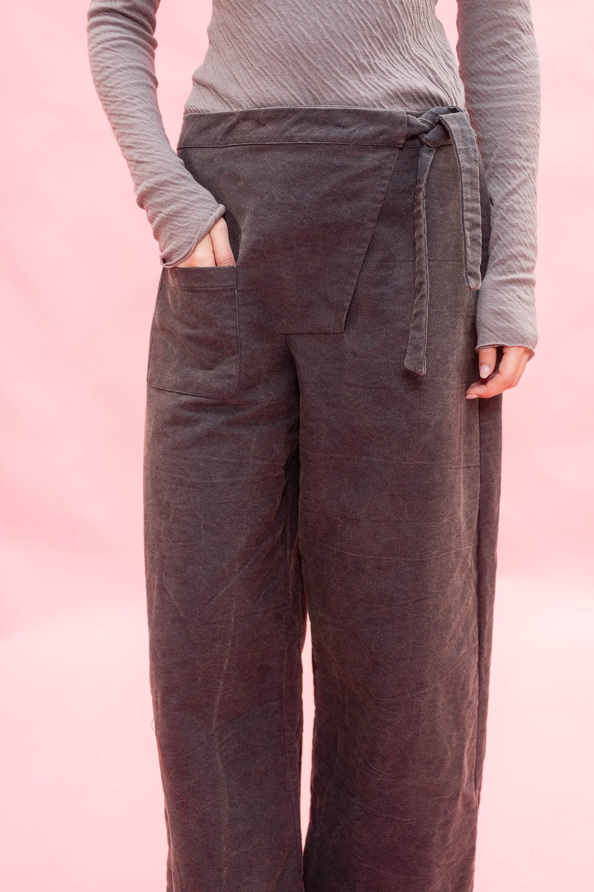 A person wearing dark na wide-leg linen pants with a tie waist and side pockets, paired with a gray ribbed long-sleeve top, set against a solid pink background.y